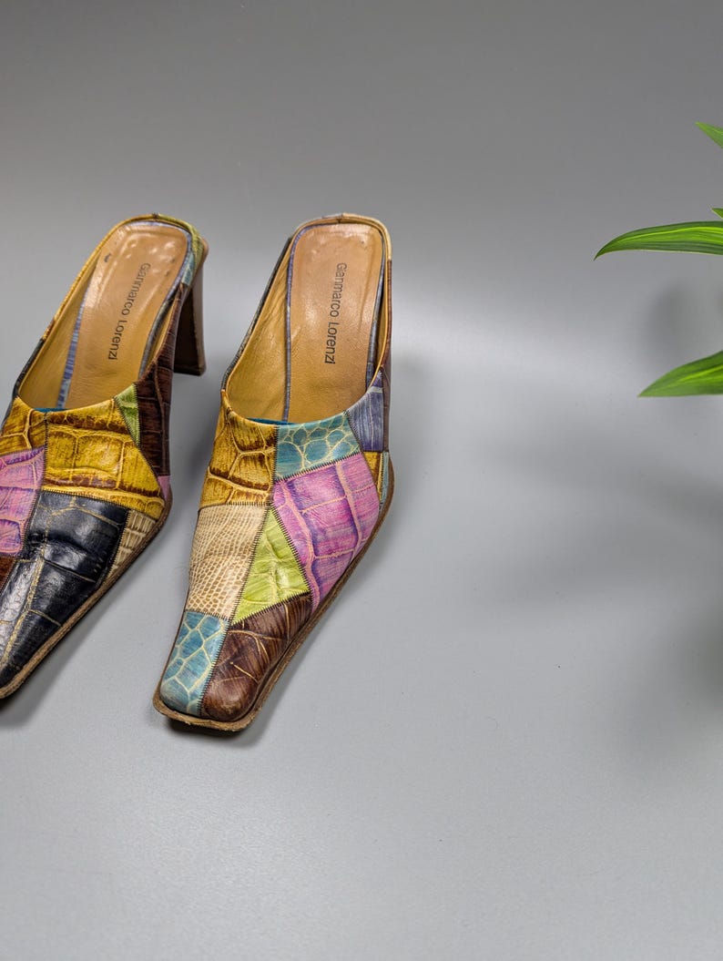 May include: A pair of patchwork leather mules with a square toe and a stacked heel. The shoes feature a variety of colors, including yellow, blue, pink, and brown. The brand name "Gennaro Lorenzi" is visible on the inner sole.