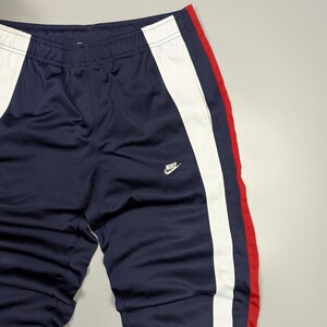 May include: Navy blue tracksuit bottoms with white and red stripes. The trousers have a white stripe down one side and a red stripe on the other. The Nike logo is visible on the front.