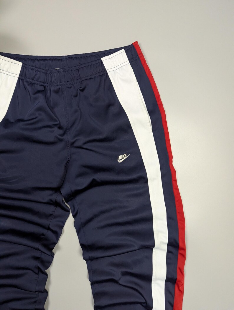 May include: Navy blue tracksuit bottoms with white and red stripes. The trousers have a white stripe down one side and a red stripe on the other. The Nike logo is visible on the front.
