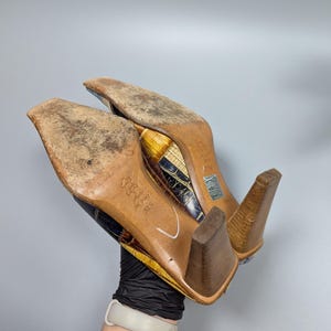 May include: A pair of heeled shoes with a patterned design in shades of brown, yellow, and blue. The soles are worn, and the heels are a light brown color. The shoes are held up, showing the bottom.