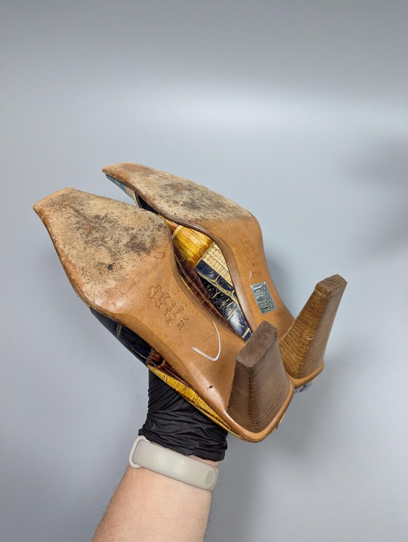 May include: A pair of heeled shoes with a patterned design in shades of brown, yellow, and blue. The soles are worn, and the heels are a light brown color. The shoes are held up, showing the bottom.