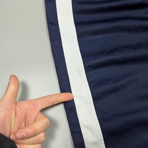May include: Navy blue athletic trousers with a broad white stripe down the side. A thin red stripe is visible near the top. The trousers are made of a smooth, possibly nylon, material.