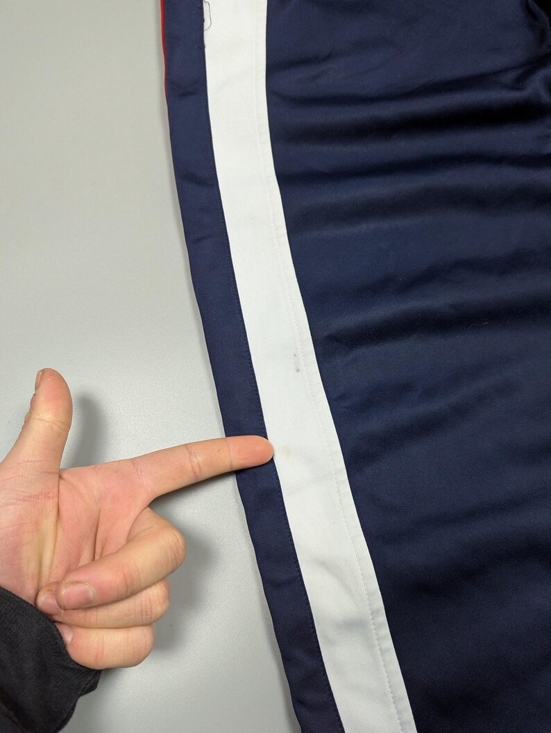 May include: Navy blue athletic trousers with a broad white stripe down the side. A thin red stripe is visible near the top. The trousers are made of a smooth, possibly nylon, material.