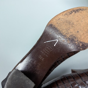 Może przedstawiać: Zbliżenie na podeszwę brązowego sk&oacute;rzanego buta. But jest oznaczony napisem "Made in Italy" i rozmiarem 38 1/2. Obcas jest w kolorze ciemnobrązowym. But ma teksturowany wz&oacute;r przypominający krokodyla.
