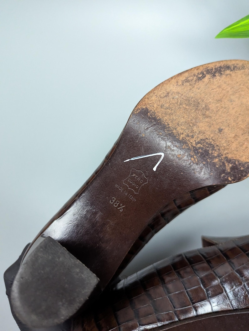 Może przedstawiać: Zbliżenie na podeszwę brązowego sk&oacute;rzanego buta. But jest oznaczony napisem "Made in Italy" i rozmiarem 38 1/2. Obcas jest w kolorze ciemnobrązowym. But ma teksturowany wz&oacute;r przypominający krokodyla.