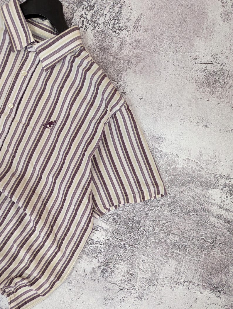 K&ouml;nnte beinhalten: Kurz&auml;rmeliges Button-Down-Hemd mit vertikalen Streifen in Braun-, Beige- und Graut&ouml;nen. Das Hemd hat ein kleines K&auml;nguru-Logo auf der Brust. Das Hemd h&auml;ngt an einem schwarzen Kleiderb&uuml;gel.