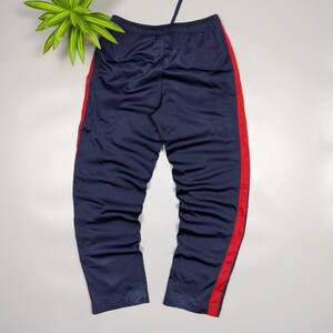 May include: Navy blue tracksuit bottoms with a red stripe down the side. The trousers have a drawstring waist and are made of a smooth material. The bottoms are displayed on a white surface.