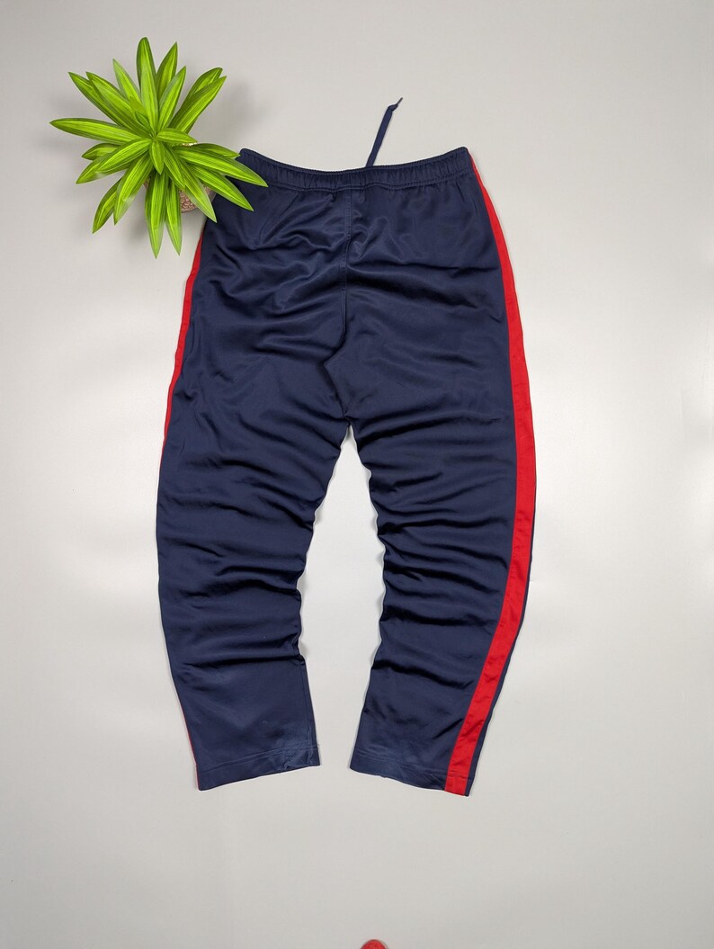 May include: Navy blue tracksuit bottoms with a red stripe down the side. The trousers have a drawstring waist and are made of a smooth material. The bottoms are displayed on a white surface.