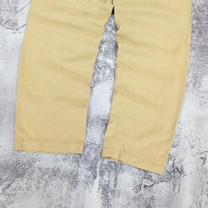 May include: A pair of solid light tan trousers featuring a straight-leg cut, a clean hemline, and a simple waistband. The fabric has a subtle texture and a consistent matte finish throughout the garment, showing the lower portion of the legs.