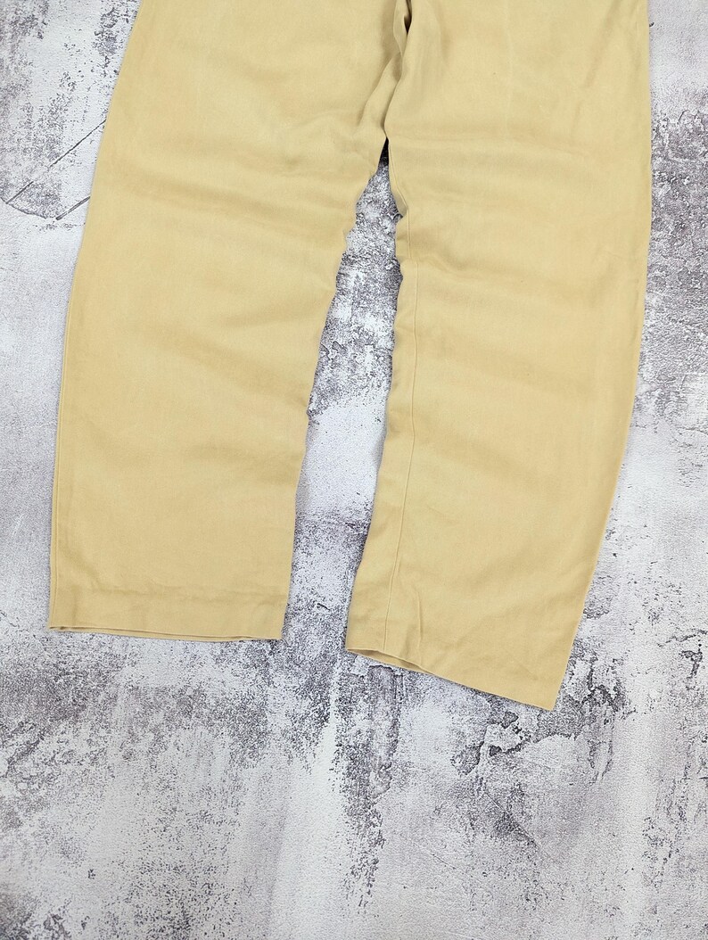 May include: A pair of solid light tan trousers featuring a straight-leg cut, a clean hemline, and a simple waistband. The fabric has a subtle texture and a consistent matte finish throughout the garment, showing the lower portion of the legs.