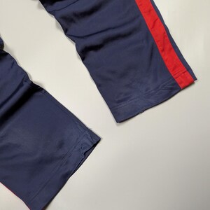 May include: Navy blue athletic trousers with a bold red stripe running down the side. The material appears smooth, possibly synthetic, and shows signs of light wear. The image focuses on the lower sections of the trousers.