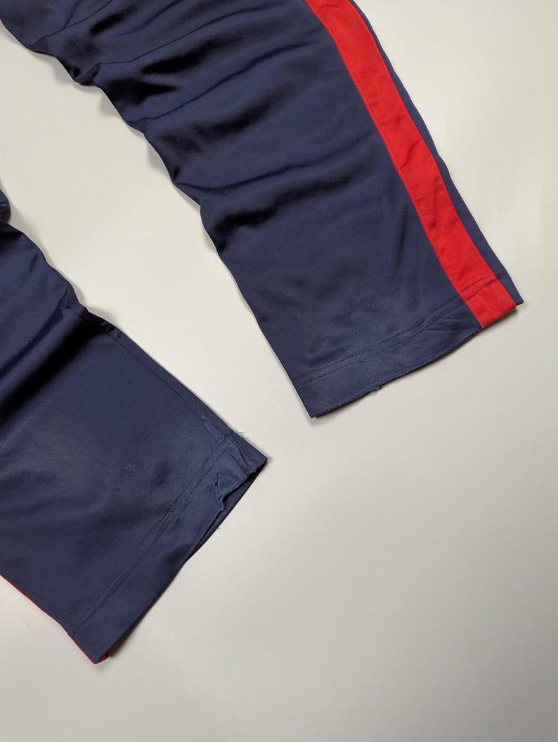 May include: Navy blue athletic trousers with a bold red stripe running down the side. The material appears smooth, possibly synthetic, and shows signs of light wear. The image focuses on the lower sections of the trousers.