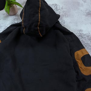 Pode incluir: Uma camisola com capuz preta com debrum de contraste castanho ao longo das costuras do capuz e grandes remendos castanhos nos cotovelos das mangas. A pe&ccedil;a &eacute; feita de um tecido escuro com uma textura vis&iacute;vel e inclui um capuz central.