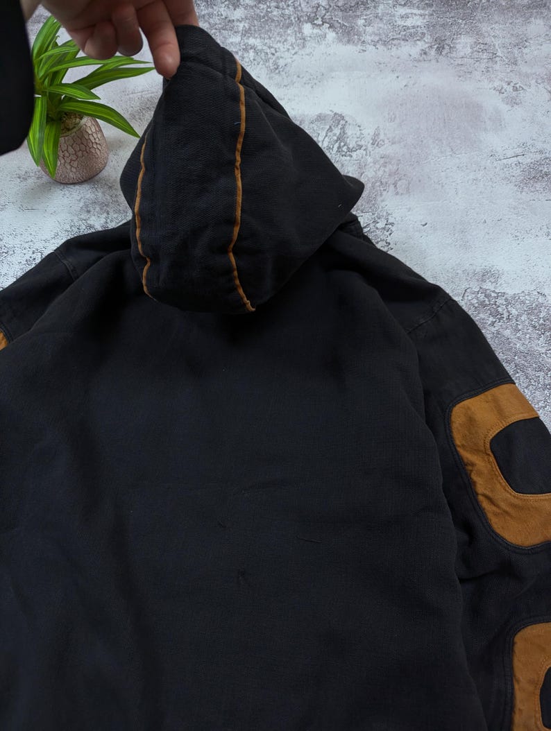 Pode incluir: Uma camisola com capuz preta com debrum de contraste castanho ao longo das costuras do capuz e grandes remendos castanhos nos cotovelos das mangas. A pe&ccedil;a &eacute; feita de um tecido escuro com uma textura vis&iacute;vel e inclui um capuz central.