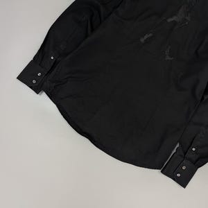 Puede incluir: Camisa negra de manga larga con pu&ntilde;os abotonados. Presenta un peque&ntilde;o parche con la bandera austriaca en la manga superior y un dise&ntilde;o de silueta oscura en la espalda. La camisa est&aacute; hecha de una tela lisa y de color s&oacute;lido.