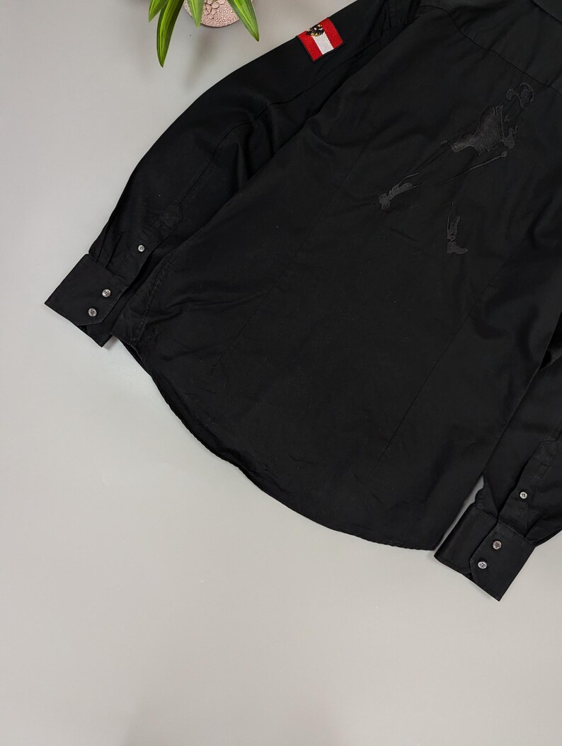 Puede incluir: Camisa negra de manga larga con pu&ntilde;os abotonados. Presenta un peque&ntilde;o parche con la bandera austriaca en la manga superior y un dise&ntilde;o de silueta oscura en la espalda. La camisa est&aacute; hecha de una tela lisa y de color s&oacute;lido.