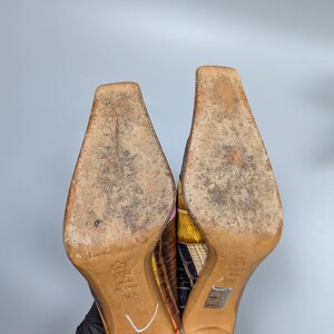 May include: A pair of colorful, patterned high-heeled shoes. The soles are light brown with wear marks. The heels are a light brown wood. The uppers feature a patchwork design with various colors and textures.