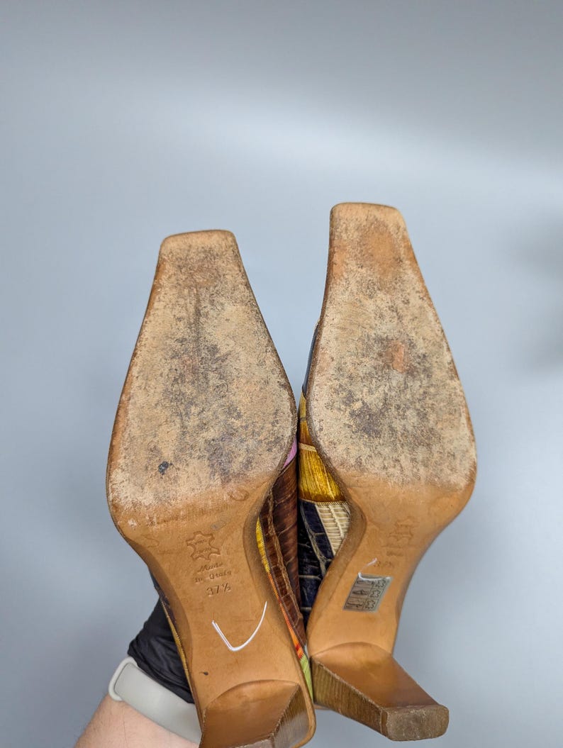 May include: A pair of colorful, patterned high-heeled shoes. The soles are light brown with wear marks. The heels are a light brown wood. The uppers feature a patchwork design with various colors and textures.
