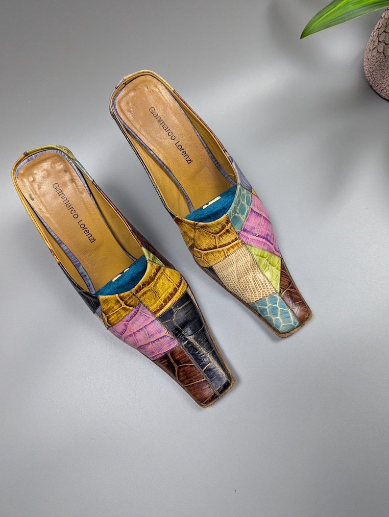 May include: A pair of Gianmarco Lorenzi patchwork leather mules. The shoes feature a variety of colors including pink, yellow, brown, and blue. The heels are low and the shoes have a pointed toe.