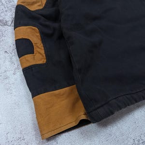 Pode incluir: Um casaco preto com remendos contrastantes em tom castanho na manga e um punho largo da mesma cor no pulso. A pe&ccedil;a &eacute; feita de um tecido resistente com costuras vis&iacute;veis ao longo das jun&ccedil;&otilde;es e um design estruturado e dur&aacute;vel.