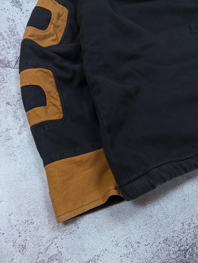 Pode incluir: Um casaco preto com remendos contrastantes em tom castanho na manga e um punho largo da mesma cor no pulso. A pe&ccedil;a &eacute; feita de um tecido resistente com costuras vis&iacute;veis ao longo das jun&ccedil;&otilde;es e um design estruturado e dur&aacute;vel.