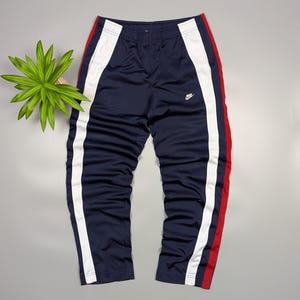 May include: Navy blue track pants with white and red stripes down the sides. The trousers feature an elasticated waistband and a small Nike logo. The trousers are made of a smooth, athletic material.