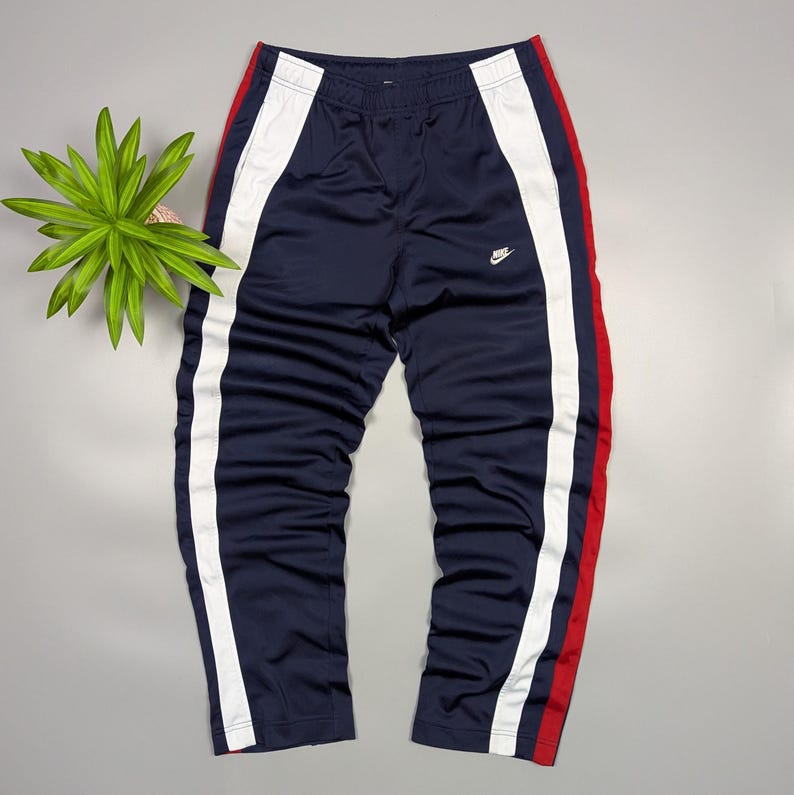 May include: Navy blue track pants with white and red stripes down the sides. The trousers feature an elasticated waistband and a small Nike logo. The trousers are made of a smooth, athletic material.