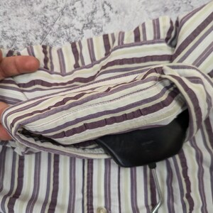 K&ouml;nnte beinhalten: Nahaufnahme eines gestreiften Button-Down-Hemds in Lila, Wei&szlig; und Hellgr&uuml;n. Das Hemd h&auml;ngt an einem schwarzen Kleiderb&uuml;gel. Der Stoff hat vertikale Streifen und einen Knopfverschluss.