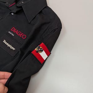 Puede incluir: Camisa negra con botones con el texto "DIAGEO AUSTRIA" en rojo. La camisa presenta un peque&ntilde;o parche de la bandera austriaca y la palabra "Teamplayer". La camisa est&aacute; colgada en una percha negra.