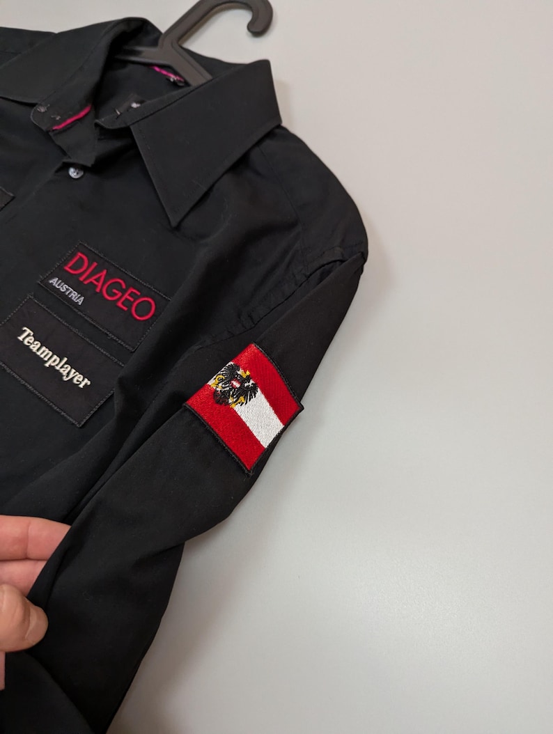 Puede incluir: Camisa negra con botones con el texto "DIAGEO AUSTRIA" en rojo. La camisa presenta un peque&ntilde;o parche de la bandera austriaca y la palabra "Teamplayer". La camisa est&aacute; colgada en una percha negra.