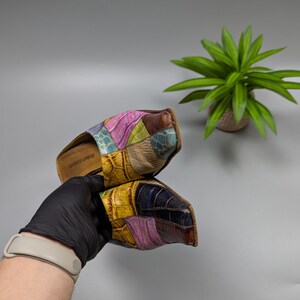 May include: A pair of colorful patchwork leather slides. The slides feature a mix of textures and colors, including pink, yellow, blue, and brown. The sole is a light brown color. The slides are held by a gloved hand.