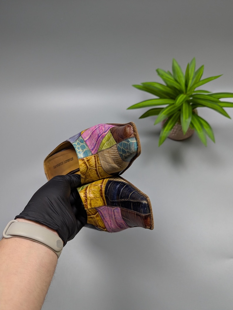 May include: A pair of colorful patchwork leather slides. The slides feature a mix of textures and colors, including pink, yellow, blue, and brown. The sole is a light brown color. The slides are held by a gloved hand.