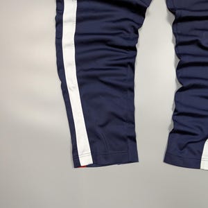 May include: Navy blue tracksuit bottoms with a broad white stripe down the side. The trousers have a straight leg design and a red accent at the hem. The fabric appears to be a smooth, synthetic material.