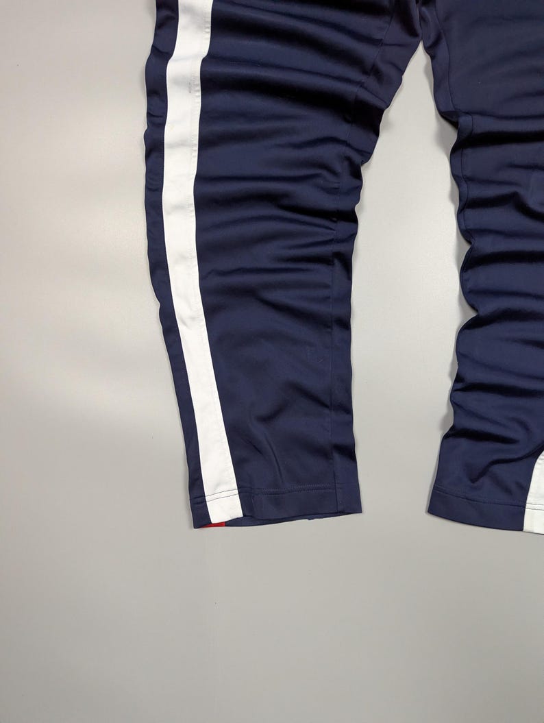 May include: Navy blue tracksuit bottoms with a broad white stripe down the side. The trousers have a straight leg design and a red accent at the hem. The fabric appears to be a smooth, synthetic material.