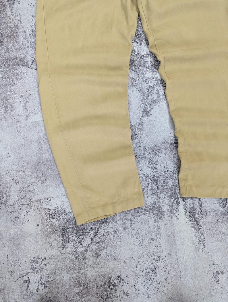 May include: A pair of light tan trousers featuring a straight-leg cut with visible vertical seam detailing running down the front of each leg. The fabric has a smooth, matte finish and the garment is presented in a simple, clean design.