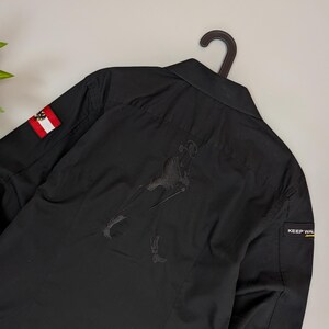 Puede incluir: Camisa negra abotonada con una figura bordada en la espalda. Incluye un parche de la bandera austriaca en la manga izquierda y una etiqueta "KEEP WAL" en la manga derecha. La camisa est&aacute; colgada de una percha negra.