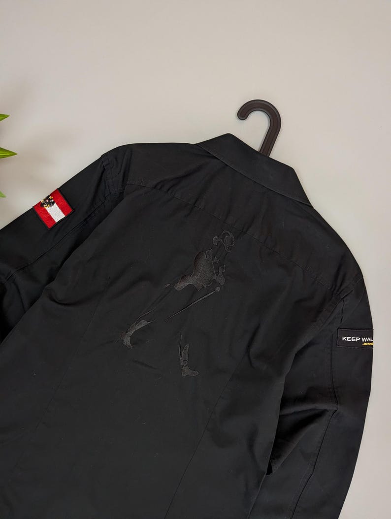 Puede incluir: Camisa negra abotonada con una figura bordada en la espalda. Incluye un parche de la bandera austriaca en la manga izquierda y una etiqueta "KEEP WAL" en la manga derecha. La camisa est&aacute; colgada de una percha negra.
