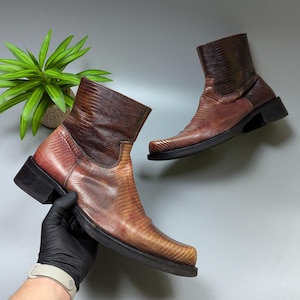 以下が含まれることがあります： 茶色の革製アンクルブーツのペア。爬虫類のような質感のある模様が特徴です。ブーツはスクエアトゥで、短い黒いヒールが付いています。片方のブーツは手袋をした手で持たれ、もう片方は空中に浮いています。背景には緑の植物があります。