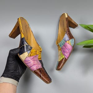 May include: A pair of patchwork leather mules with wooden block heels. The shoes feature a variety of colors including brown, yellow, pink, blue, and green. The brand name "Gianmarco Lorenzi" is visible on the inner sole.