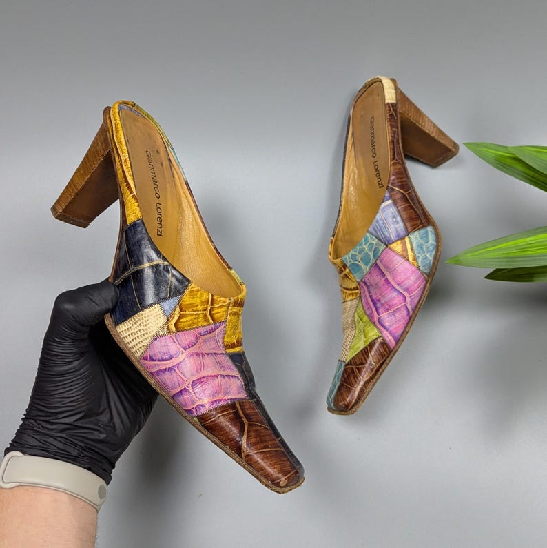 May include: A pair of patchwork leather mules with wooden block heels. The shoes feature a variety of colors including brown, yellow, pink, blue, and green. The brand name "Gianmarco Lorenzi" is visible on the inner sole.