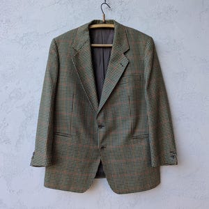 May include: A houndstooth blazer in shades of green, brown, and black hangs on a wooden hanger. The jacket has a classic two-button closure, notched lapels, and two front pockets. The lining is a solid dark gray color.