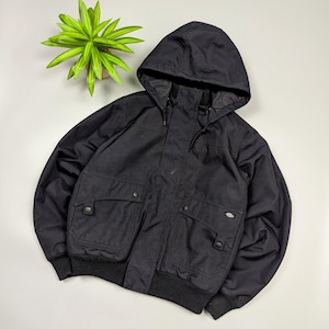 May include: Black hooded jacket with multiple front pockets and a ribbed hem. The jacket features a Dickies logo patch on the front and a full-length zipper closure. The jacket has a hood and is made of a durable fabric.