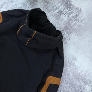Pode incluir: Uma camisola com capuz preta com remendos nos cotovelos em imita&ccedil;&atilde;o de camur&ccedil;a castanha e detalhes de debrum castanho ao longo das costuras do capuz. A pe&ccedil;a &eacute; feita de um tecido escuro e espesso com um forro interior vis&iacute;vel.