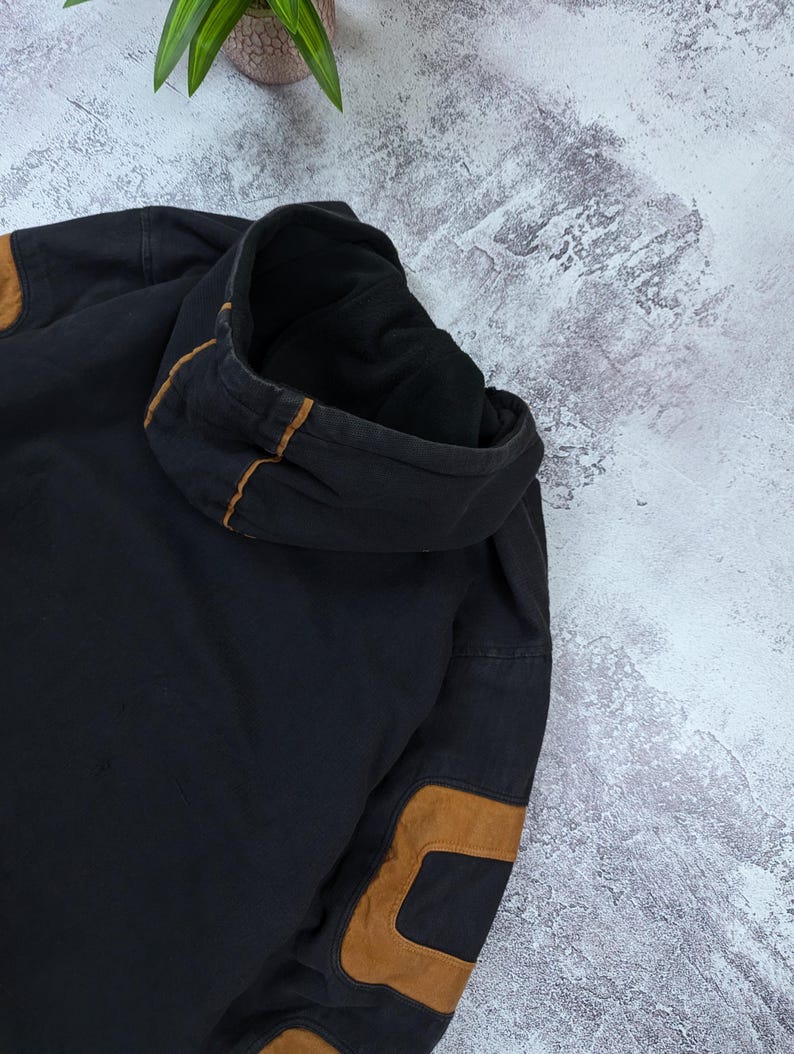 Pode incluir: Uma camisola com capuz preta com remendos nos cotovelos em imita&ccedil;&atilde;o de camur&ccedil;a castanha e detalhes de debrum castanho ao longo das costuras do capuz. A pe&ccedil;a &eacute; feita de um tecido escuro e espesso com um forro interior vis&iacute;vel.