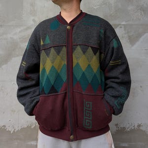May include: A gray bomber jacket with a geometric pattern in teal, yellow, and burgundy. The jacket features a burgundy trim, pockets, and a button closure. The design includes diamond shapes and other abstract elements.
