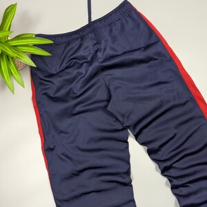 May include: Navy blue tracksuit bottoms with a red stripe down the side. The trousers have a drawstring at the waist. The fabric has a slight sheen. The trousers are folded and laid flat.