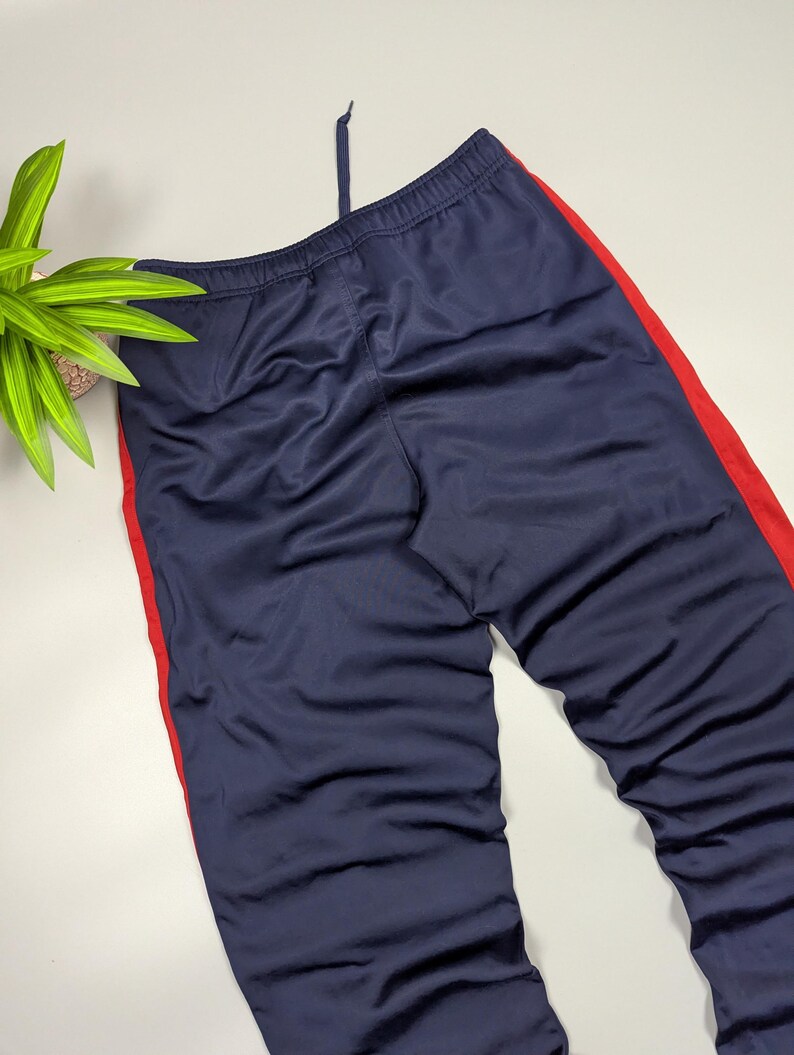 May include: Navy blue tracksuit bottoms with a red stripe down the side. The trousers have a drawstring at the waist. The fabric has a slight sheen. The trousers are folded and laid flat.