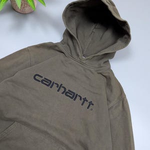 Puede incluir: Una sudadera con capucha Carhartt verde oliva descolorida con capucha con cord&oacute;n y bolsillo de canguro. El nombre de la marca est&aacute; bordado en gris oscuro en el pecho. La sudadera est&aacute; hecha de un material suave y desgastado.