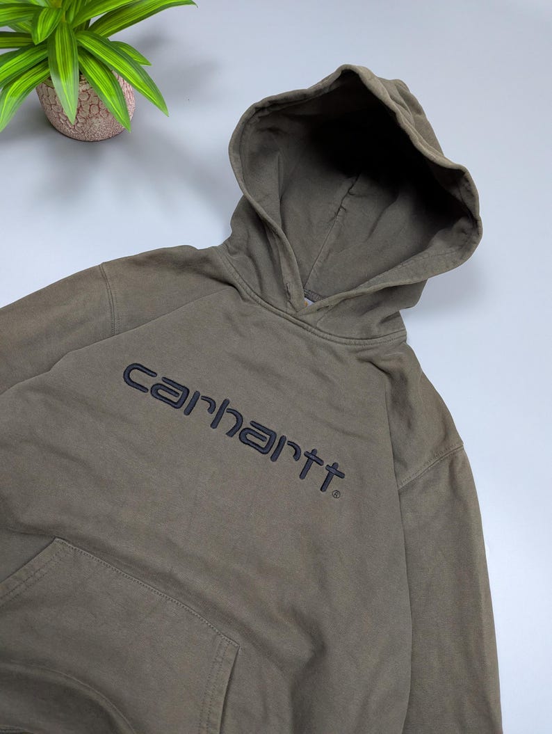 Puede incluir: Una sudadera con capucha Carhartt verde oliva descolorida con capucha con cord&oacute;n y bolsillo de canguro. El nombre de la marca est&aacute; bordado en gris oscuro en el pecho. La sudadera est&aacute; hecha de un material suave y desgastado.