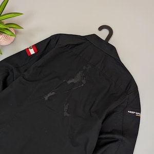 Puede incluir: Camisa negra con botones con un peque&ntilde;o parche de la bandera austriaca en la manga. La parte trasera presenta una gran figura negra estilizada. Una peque&ntilde;a etiqueta en la manga dice "KEEP WAL". La camisa est&aacute; colgada en una percha negra.
